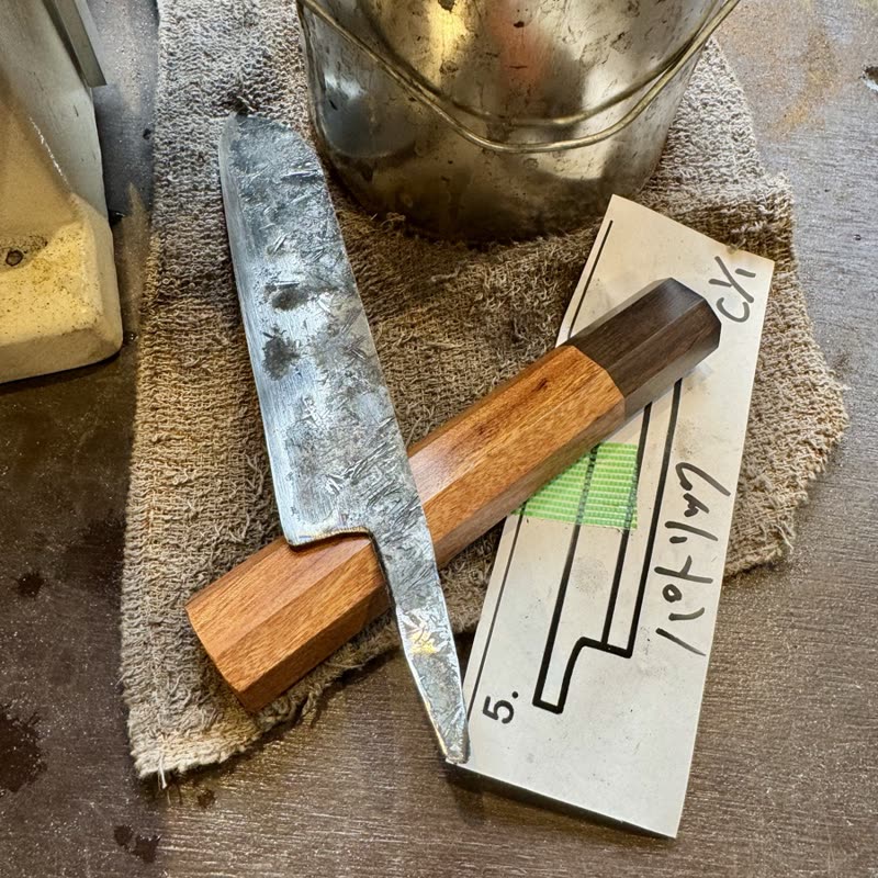A hand-forged knife with a wooden handle beside its paper template